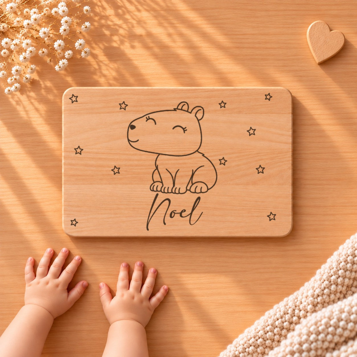 Personalisiertes Frühstücksbrettchen aus Buche mit einem Capybara, Sternen und Namen Noel, daneben Babyhände