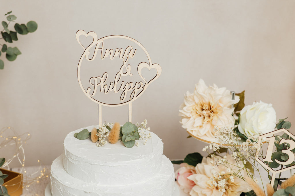 Runder personalisierter Caketopper zur Hochzeit neben Blumen und Tischnummer